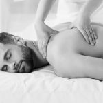 Younger man with beard lying supine with with eyes closed while massage therapist hands work thumbs behind the scapula