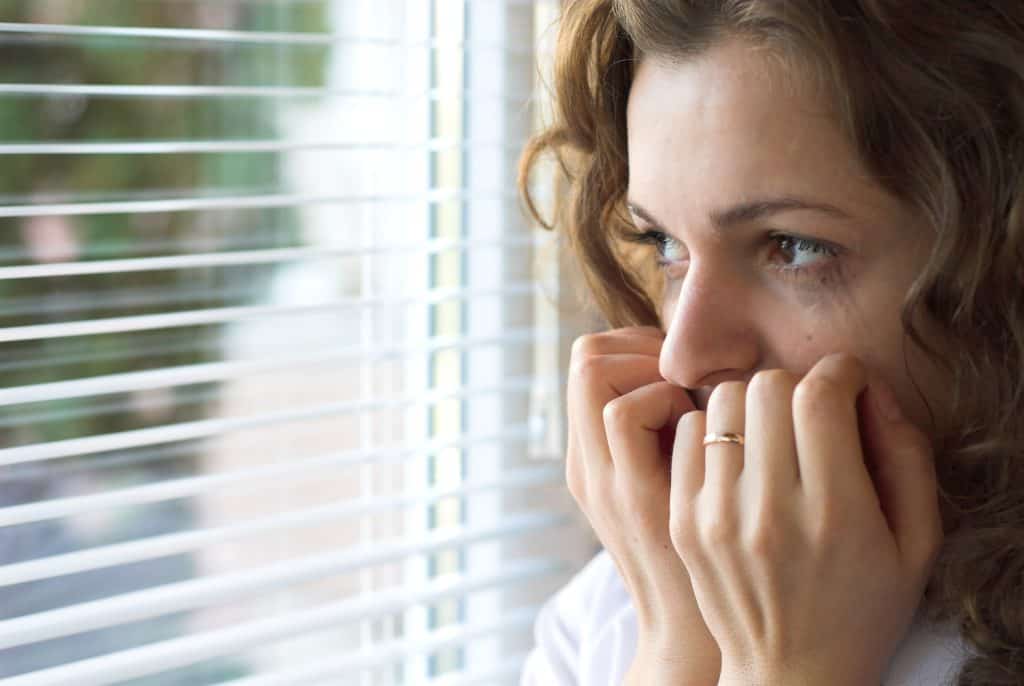 Young woman staring out window through blinds in fear