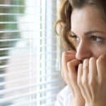 Young woman staring out window through blinds in fear